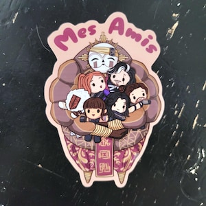 Puede incluir: Una pegatina con un grupo de personajes de dibujos animados abrazándose. La pegatina tiene un borde rosa con las palabras "Mes Amis". Los personajes están en un diseño marrón y morado.