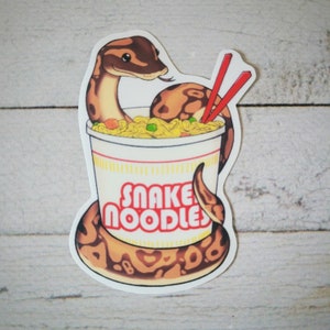 Puede incluir: Una pegatina con una serpiente de dibujos animados en una taza de fideos. La serpiente es marrón y canela, con una cara sonriente. La taza es blanca con las palabras "Snake Noodles" en rojo. Palillos chinos rojos están en los fideos.