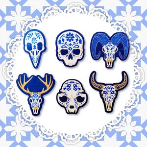 May include: A collection of six decorative skull and animal skull designs in blue and white. Each design features intricate floral patterns and gold accents. The skulls are arranged on a white, scalloped doily against a blue and white patterned background.