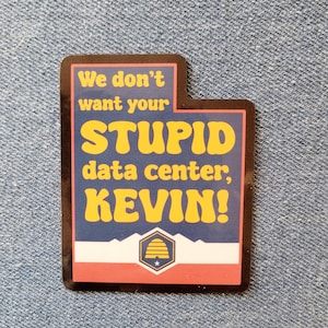 May include: A rectangular sticker shaped like the state of Utah with a dark blue background and yellow text that reads, We don't want your STUPID data center, KEVIN! Below the text is a white mountain silhouette and a beehive emblem in a blue hexagon.