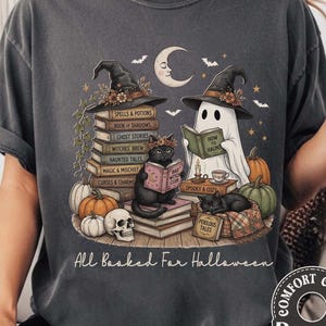 May include: A charcoal gray t-shirt featuring a graphic of a ghost and two black cats wearing witch hats, surrounded by stacks of books, pumpkins, a skull, and a crescent moon. The text "All Booked For Halloween" is printed at the bottom.