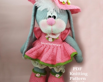 Puede incluir: Una muñeca de conejo tejida de color gris que lleva un vestido rosa con dobladillo verde, zapatos rosas con pequeñas flores blancas y un sombrero de sol rosa a juego adornado con detalles florales blancos y verdes. Aparece el texto SavushkaDesigns y PDF Knitting Pattern Knit Flat.