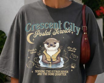 May include: A charcoal gray oversized t-shirt featuring a graphic of an otter wearing a life vest. The text reads Crescent City Postal Service, Serving the Istros River and the Bone Quarter, with small illustrations of a moon, stars, and envelopes.