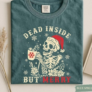 May include: A blue-green crew neck t-shirt featuring a graphic of a skeleton wearing a red Santa hat and holding a mug with a candy cane. The text reads DEAD INSIDE BUT MERRY, surrounded by white snowflakes and poinsettia flower illustrations.