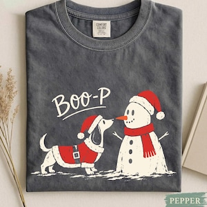 May include: A charcoal gray crew neck t-shirt featuring a graphic of a dachshund wearing a red Santa hat and coat facing a snowman with a red scarf and hat. The text "BOO-P" is printed in white above the dog. The shirt has a Comfort Colors label.
