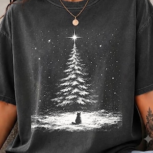 May include: A charcoal gray short-sleeve t-shirt featuring a white graphic print of a snow-covered pine tree with a bright star at the top and a small cat sitting in the snow at the base, surrounded by falling snowflakes in a winter scene.