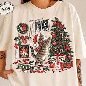 May include: An ivory t-shirt featuring a graphic of a tabby cat wearing a Santa hat and scarf, decorating a Christmas tree. The scene includes a fireplace with stockings, a wreath, wrapped gifts, and the text Comfort Colors Ivory.