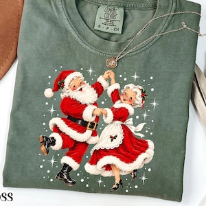 May include: A moss green crewneck t-shirt featuring a graphic of Santa Claus and Mrs. Claus dancing together. The graphic shows them in red and white holiday attire surrounded by white starbursts and dots. The shirt has a Comfort Colors brand label.