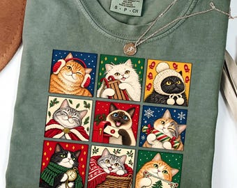May include: A moss green crew neck t-shirt featuring a grid of nine illustrated cats in winter attire. The graphic includes cats with earmuffs, scarves, and holiday items like a mailbox and a basket. The shirt has a Comfort Colors brand label.
