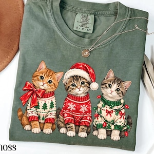 May include: A moss green t-shirt featuring a graphic of three kittens. The left kitten wears a sweater with green trees and a red bow. The center kitten wears a red sweater with snowflakes and a Santa hat. The right kitten wears a sweater with green trees and red bows, wrapped in colorful Christmas lights.