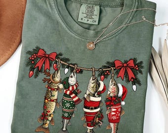 May include: A moss green t-shirt featuring a graphic of four fish hanging from a string decorated with pine branches, red bows, and Christmas lights. Each fish wears festive holiday attire, including a Santa hat, scarf, and sweater.