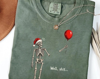 May include: A moss green t-shirt featuring a graphic of a skeleton wearing a red Santa hat and holding a red balloon. The text "Well, shit..." is printed below the skeleton. The shirt has a Comfort Colors brand label at the inner neckline.