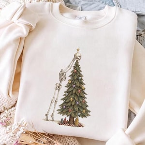 May include: A cream-colored crewneck sweatshirt featuring a graphic print of a human skeleton placing a star on top of a green Christmas tree. The tree is decorated with small, colorful skull ornaments and string lights, with a gift box at the base.