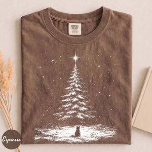May include: A brown short-sleeve t-shirt featuring a white graphic of a snow-covered pine tree with a star at the top and a small cat sitting at the base. The shirt has a crew neckline and a Comfort Colors brand label on the inside collar.