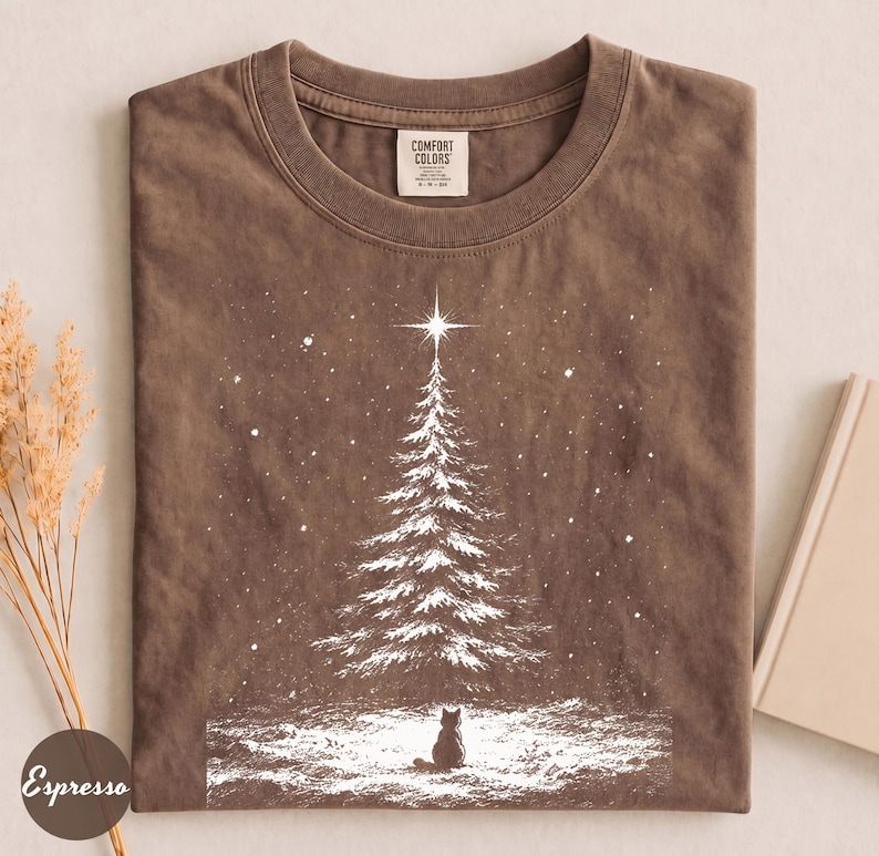 May include: A brown short-sleeve t-shirt featuring a white graphic of a snow-covered pine tree with a star at the top and a small cat sitting at the base. The shirt has a crew neckline and a Comfort Colors brand label on the inside collar.