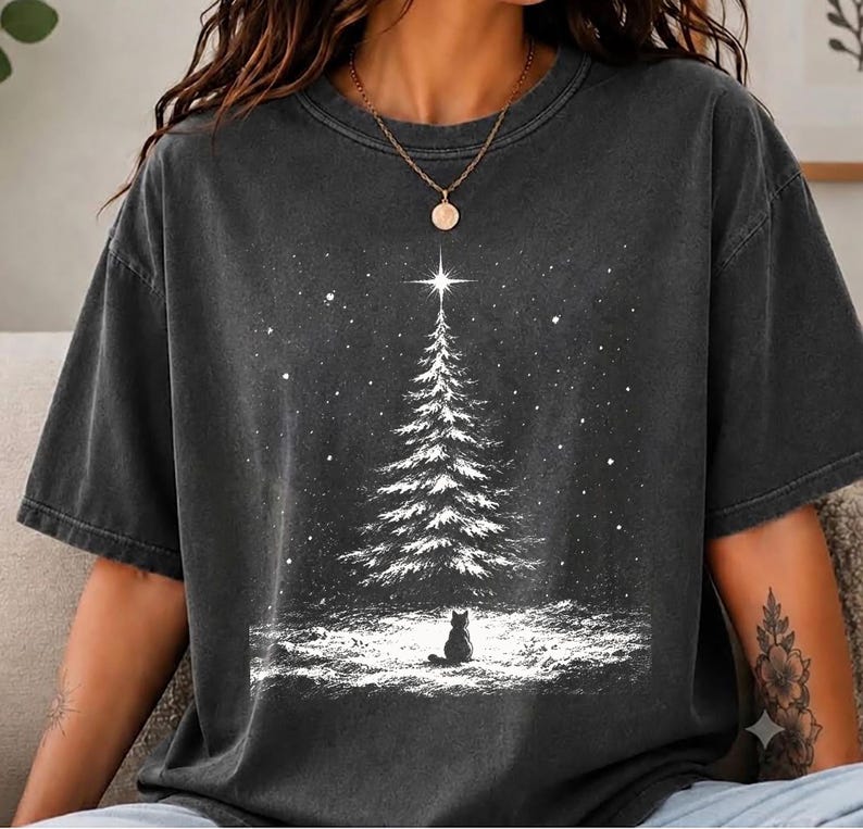 May include: A charcoal gray short-sleeve t-shirt featuring a white graphic print of a snow-covered pine tree with a bright star at the top and a small cat sitting in the snow at the base, surrounded by falling snowflakes in a winter scene.