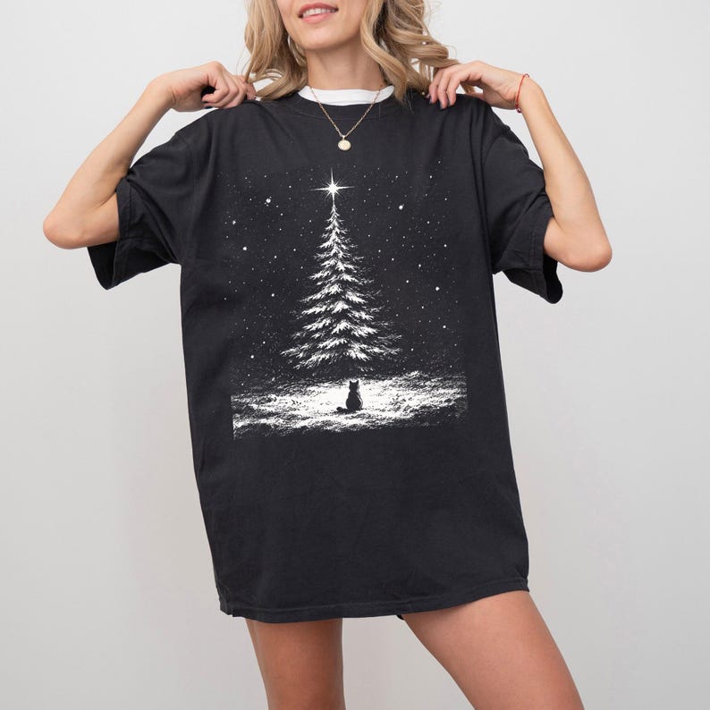 May include: A black short-sleeved t-shirt featuring a white graphic print of a snow-covered pine tree topped with a bright star, with a small cat sitting in the snow below. The design includes scattered white dots representing falling snow.