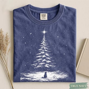 May include: A navy blue crew neck t-shirt featuring a white graphic print of a snow-covered pine tree topped with a bright star and a small cat sitting in the snow at the base. The inner neck label displays the text Comfort Colors.