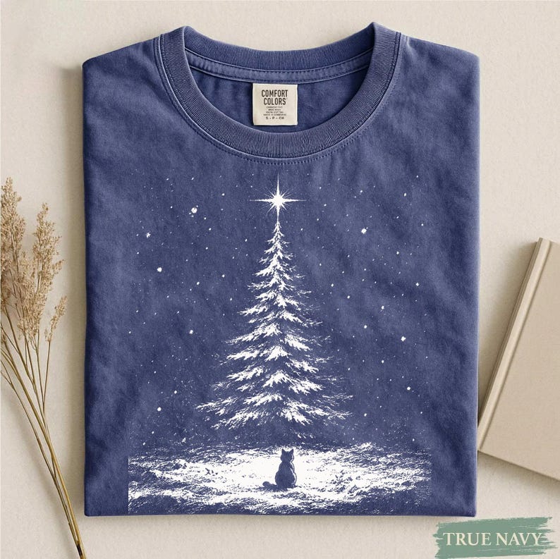 May include: A navy blue crew neck t-shirt featuring a white graphic print of a snow-covered pine tree topped with a bright star and a small cat sitting in the snow at the base. The inner neck label displays the text Comfort Colors.
