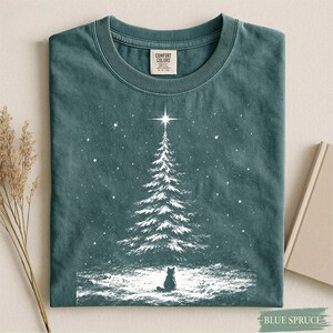 May include: A dark teal crew neck t-shirt featuring a white graphic print of a snow-covered pine tree topped with a bright star. A small black cat sits at the base of the tree in the snow. The shirt has a Comfort Colors brand label at the neck.