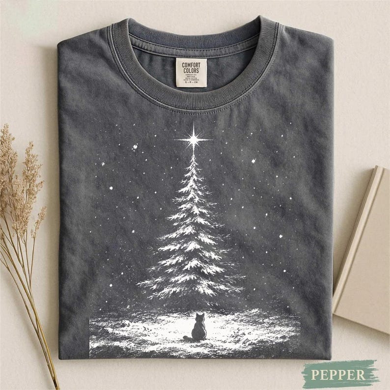 May include: A charcoal gray crew neck t-shirt featuring a white graphic print of a snow-covered pine tree topped with a bright star, with a small cat silhouette sitting in the foreground amidst falling snowflakes and a snowy ground texture.