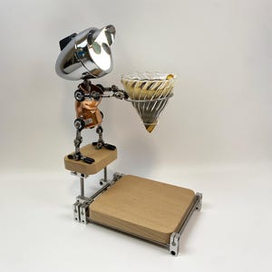 Könnte beinhalten: Ein einzigartiger Kaffeebrühständer mit Roboterdesign. Der Ständer hat einen verchromten Kopf, einen Kupfertorso und gegliederte Gliedmaßen. Er hält einen Glas-Pour-Over-Kaffeetropfer über einer Holzbasis. Der Ständer besteht aus Metall und Holz.