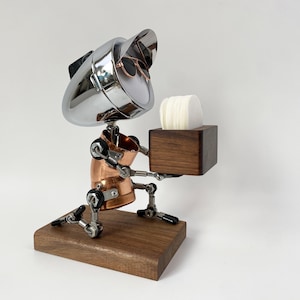 May include: A handcrafted robot sculpture made from metal, copper, and wood. The robot holds a wooden box containing white circular objects. The robot's head is a chrome-plated object with sunglasses. The sculpture is mounted on a wooden base.