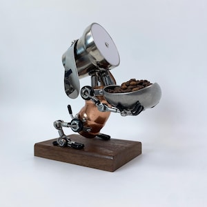 Könnte beinhalten: Handgefertigte Metallskulptur eines hundeartigen Roboters, der eine Schale mit Kaffeebohnen hält. Die Skulptur besteht aus Edelstahl, Kupfer und anderen Metallteilen, die auf einem dunklen Holzsockel montiert sind.