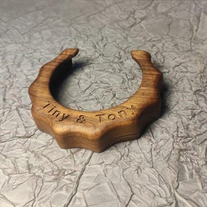 Könnte beinhalten: Ein handgefertigtes, hufeisenförmiges Objekt aus Holz mit dem eingravierten Text "Tiny & Tony". Das Objekt besteht aus hellbraunem Holz und hat einen strukturierten, gewellten Rand.