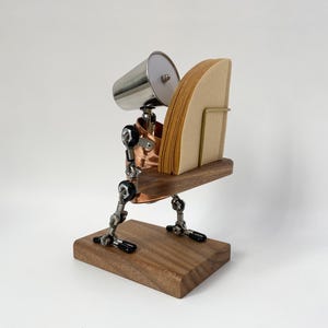Könnte beinhalten: Ein einzigartiger Kaffeefilterhalter mit Steampunk-Ästhetik. Er verfügt über einen Holzsockel, mechanische Komponenten aus Kupfer und Stahl sowie ein becherartiges Element aus Edelstahl. Der Halter ist für eine Stapel brauner Papierkaffeefilter konzipiert.