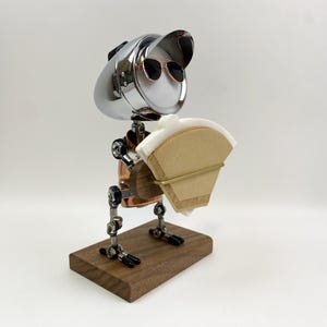 May include: A unique coffee filter holder in the shape of a robot. The robot has a chrome head with sunglasses, copper body, and metal legs. It holds a stack of paper coffee filters. The base is a rectangular wooden block.