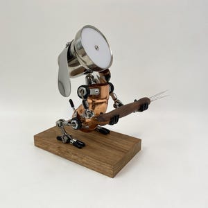Könnte beinhalten: Eine einzigartige Skulptur eines Roboterhundes aus Metall- und Kupferkomponenten. Der Roboterhund hält ein Holzobjekt mit Metallzinken. Er ist auf einem rechteckigen Holzsockel montiert. Der Roboterhund hat ein großes, rundes, weißes Licht auf dem Kopf.