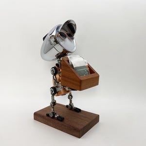 Könnte beinhalten: Ein roboterförmiger Schreibtisch-Organizer aus Chrom, Kupfer und Holz. Der Kopf verfügt über ein Chromvisier und eine kleine Sonnenbrille. Vorne befindet sich eine Holzbox zur Aufbewahrung, montiert auf einem rechteckigen Holzsockel.