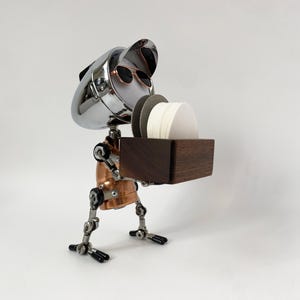 Op de afbeelding: Een robotfiguur gemaakt van metalen onderdelen, met een verchroomde kop met vizier en zonnebril, een koperkleurige romp en beweegbare ledematen. Het houdt een rechthoekige donkerhouten doos vast met een stapel ronde witte en grijze onderzetters.