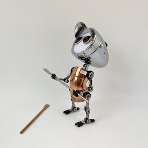 以下が含まれることがあります： クロム製の頭部、銅製の胴体、関節式の金属製の四肢を備えた、気まぐれな手作りのロボット彫刻。 ロボットはサングラスをかけ、金属製の道具を持っています。 木製のスプーンが近くにあります。 白い背景に彫刻が施されています。