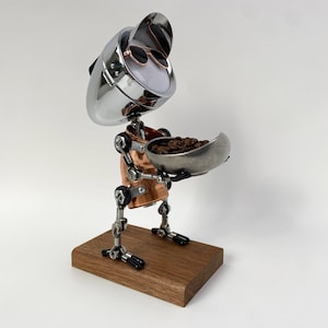 Könnte beinhalten: Eine handgefertigte Roboterskulptur aus Metall, Kupfer und Holz. Der Roboter hält eine Schale mit Kaffeebohnen. Der Kopf des Roboters ist ein verchromtes Objekt mit Sonnenbrille. Der Roboter steht auf einem Holzsockel.