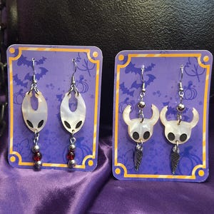 Puede incluir: Dos pares de pendientes hechos a mano exhibidos en tarjetas moradas. Un par presenta un diseño ovalado blanco con cuentas rojas, el otro tiene un diseño de personaje blanco con detalles de plumas negras. El fondo incluye un caldero y tela morada.