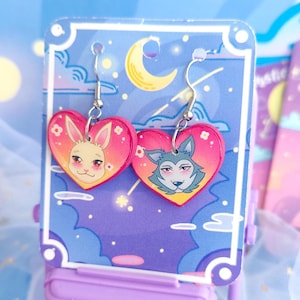May include: Heart-shaped dangle earrings featuring a bunny and a wolf character. The earrings have a pink and orange gradient background with silver-colored hooks. The characters are set against a backdrop of a blue sky with clouds and a crescent moon.