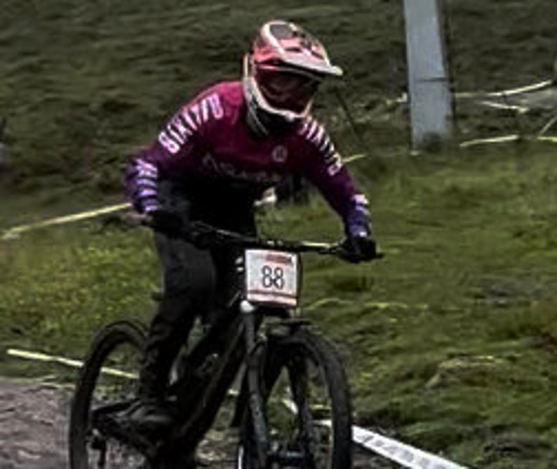 May include: A woman wearing a pink and purple long-sleeved jersey with the number 88 on her bike, riding a black mountain bike on a dirt path.