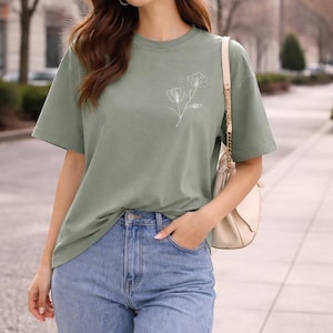 May include: A sage green short-sleeve t-shirt featuring a white line art illustration of two flowers on the left chest, paired with light blue denim jeans and a beige shoulder bag with a gold chain strap.