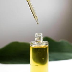 Könnte beinhalten: Eine klare Glasflasche mit einer Pipette, gefüllt mit einer gelben Flüssigkeit. Die Flasche steht auf einer weißen Oberfläche mit einem grünen Blatt dahinter.