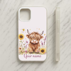 May include: A white phone case with a watercolour illustration of a Highland cow amidst sunflowers and wildflowers. The text "Your name" is written in a cursive font at the bottom. A matching white phone case is shown next to the case.
