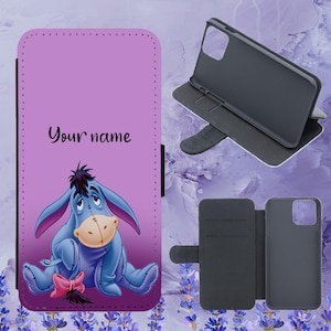 May include: A purple phone case featuring Eeyore from Winnie the Pooh, with the text "Your name". The case is open, showing the phone holder and card slots. The design includes a pink bow.