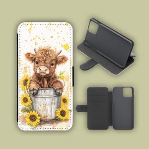 May include: A phone case with a watercolour illustration of a brown Highland cow calf in a metal bucket and yellow sunflowers. The case is white with a black interior and a magnetic closure. The image also shows the case open and the phone holder.