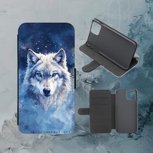 Puede incluir: Funda de teléfono con una pintura de acuarela de un lobo con ojos azules sobre un fondo azul oscuro y blanco. La funda se muestra abierta y cerrada, con un interior negro y ranuras para tarjetas.