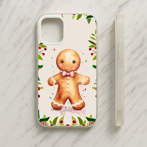 May include: A phone case featuring a watercolor illustration of a brown gingerbread person with a pink bow tie, white icing details, and rosy cheeks, framed by a border of green holly leaves and red berries on a light cream background.