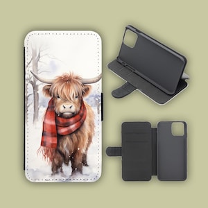 Könnte beinhalten: Eine Handyhülle mit einer Aquarellillustration einer Highland-Kuh, die einen roten Schottenkaro-Schal in einer verschneiten Winterszene trägt. Die Hülle ist weiß mit schwarzem Rand und enthält einen separaten Telefonhalter.