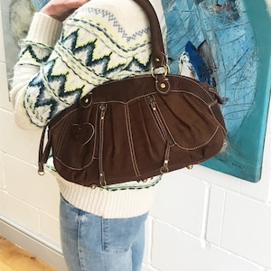 May include: A brown leather handbag with a heart-shaped detail on the front. The bag has a zipper closure and multiple pockets. The bag is being held by a person wearing a white sweater with a geometric pattern and blue jeans.