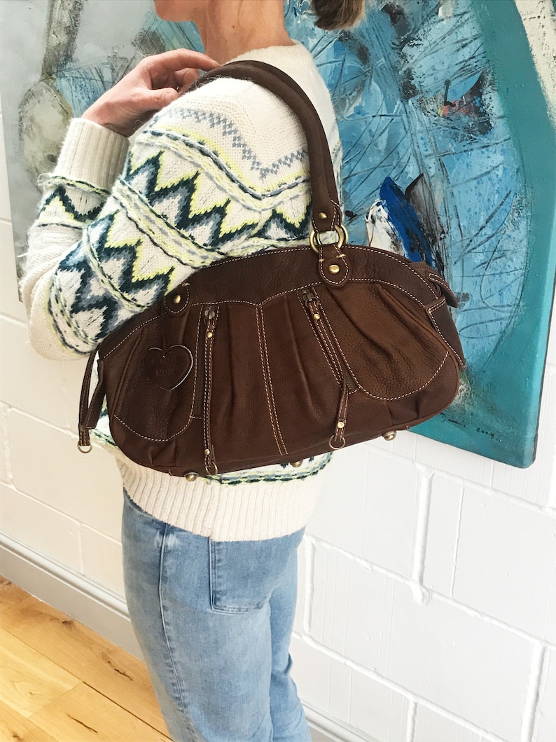 May include: A brown leather handbag with a heart-shaped detail on the front. The bag has a zipper closure and multiple pockets. The bag is being held by a person wearing a white sweater with a geometric pattern and blue jeans.