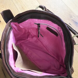 May include: Brown leather handbag with a pink fabric lining and a zippered pocket. The bag has a top handle and a zipper closure.